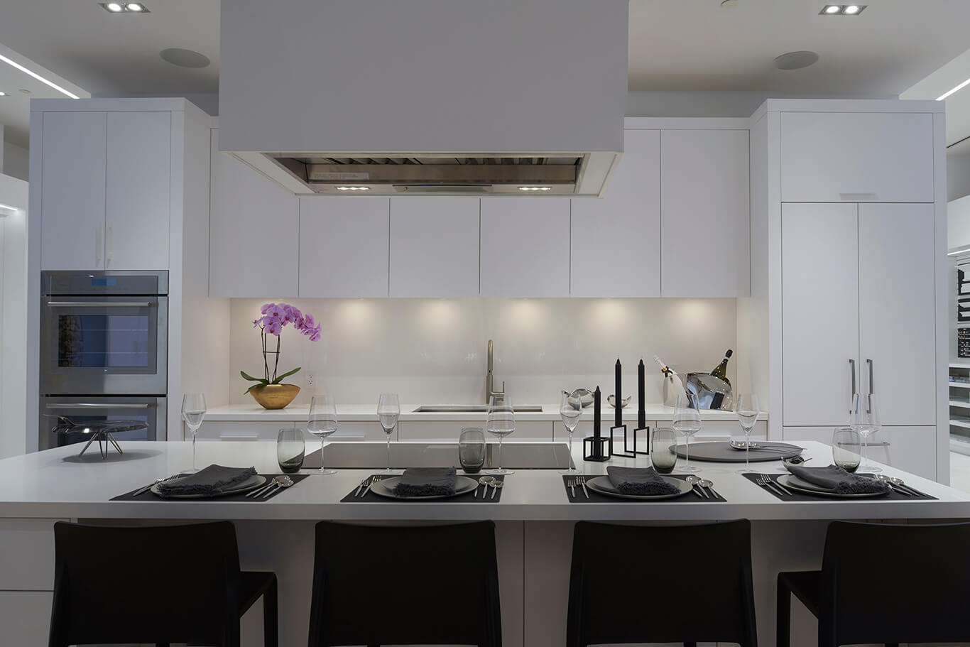 White display kitchen with a large island set for dinner, a ceiling-mounted hood and built-in wall ovens
