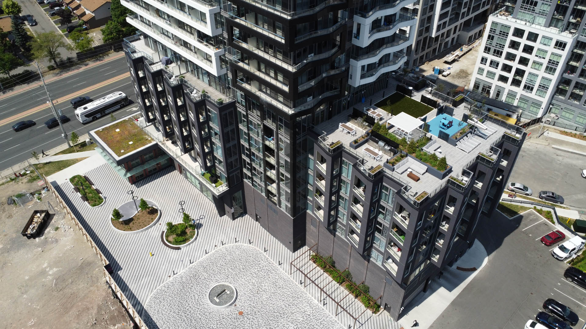 Drone view looking down on the DUO podium, its landscaped ground-level courtyard and the rooftop terrace, beside a wide road with buses